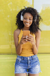 Lächelnde Frau, die ein Mobiltelefon benutzt, während sie über Kopfhörer vor gelben Jalousien Musik hört - JRVF01170