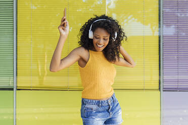 Glückliche Frau hört Musik über Kopfhörer und tanzt vor einem gelben Fenster - JRVF01175
