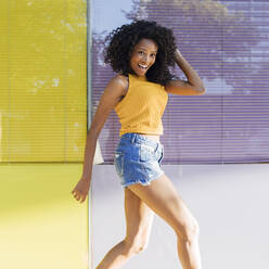 Fröhliche junge Frau mit Hand im Haar, die vor einem gelben Fenster herumspringt - JRVF01178