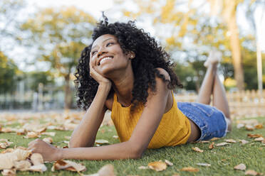 Nachdenklich lächelnde junge Frau, die wegschaut, während sie im Park im Gras liegt - JRVF01193