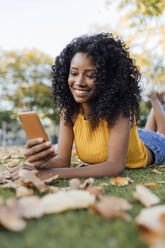 Smiling young woman text messaging through smart phone lying on grass at park - JRVF01196