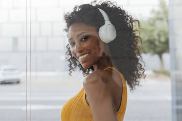 Lächelnde, lockig behaarte Frau, die über drahtlose Kopfhörer Musik hört, gesehen durch eine Glaswand - JRVF01199