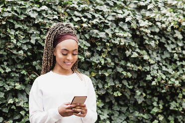 Lächelnde Frau, die eine Textnachricht über ihr Smartphone sendet, steht vor grünen Efeupflanzen - NMSF00436