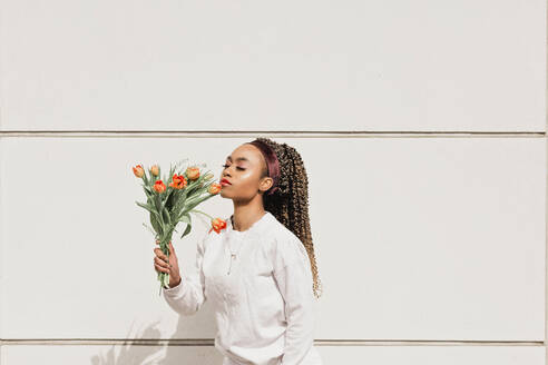 Junge Frau mit geschlossenen Augen riecht Blumen vor einer weißen Wand an einem sonnigen Tag - NMSF00444