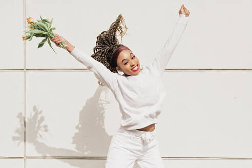 Cheerful young woman with flowers jumping in front of white wall during sunny day - NMSF00445