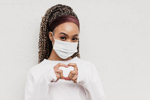 Young woman wearing protective face mask making heart shape from hands - NMSF00450