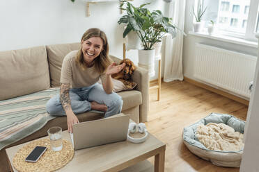 Glückliche Geschäftsfrau mit Laptop, die auf dem Sofa neben einem Hund gestikuliert - VPIF04174