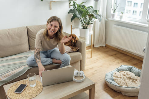 Happy businesswoman with laptop gesturing on sofa by dog at home - VPIF04174