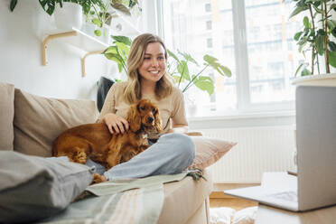 Lächelnde Geschäftsfrau, die ihren Hund während einer Videokonferenz über ihren Laptop zu Hause streichelt - VPIF04178