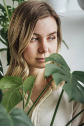 Blond woman contemplating behind plant at home - VPIF04190