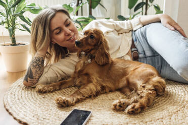 Beautiful woman looking at dog while relaxing on rug in living room - VPIF04196