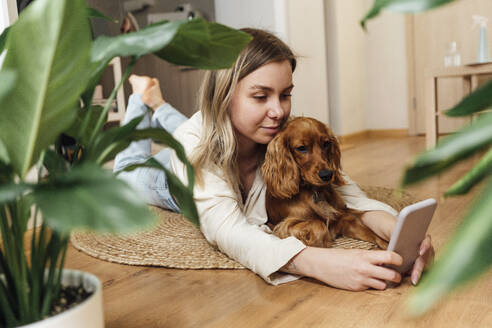 Junge Frau mit Cocker Spaniel benutzt ihr Smartphone, während sie zu Hause auf dem Boden liegt - VPIF04197