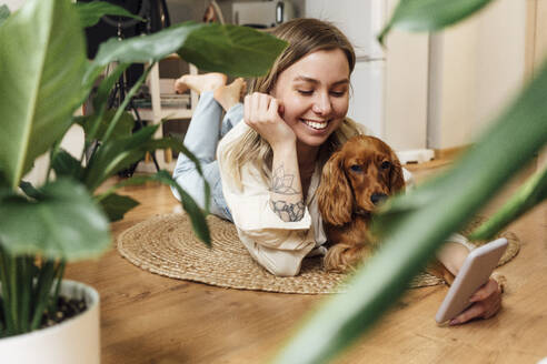 Lächelnde schöne Frau mit Hund, die ein Smartphone benutzt, während sie zu Hause auf dem Teppich liegt - VPIF04199