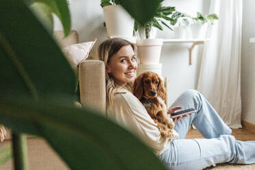 Lächelnde junge Frau mit Hund hält Smartphone zu Hause - VPIF04213