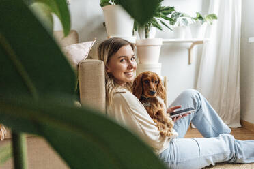 Smiling young woman with dog holding smart phone at home - VPIF04213