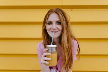 Smiling woman holding smoothie in front of yellow wall - TCEF01958