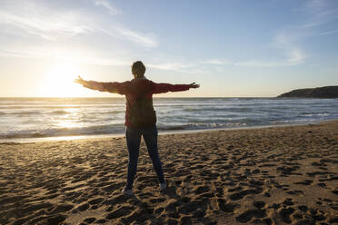 Woman standing with arms outstretched at beach during sunset - WPEF04925