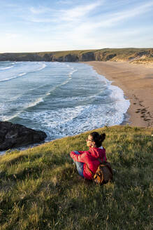 Frau mit Rucksack sitzt auf Gras in der Nähe von Strand bei Sonnenuntergang - WPEF04927