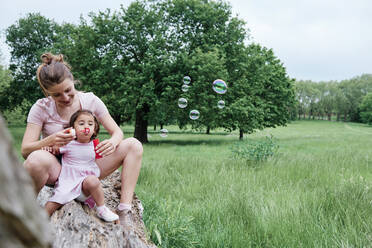 Daughter blowing bubbles with mother sitting on log in park - ASGF00687