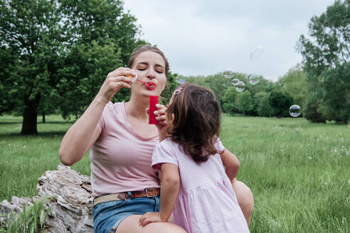 Playful mother blowing bubbles with daughter in park - ASGF00688