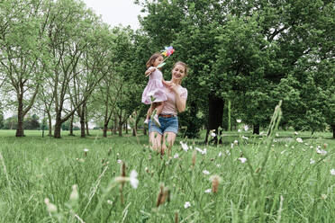 Glückliche Mutter, die ihre Tochter mit einem Windrad trägt, während sie im Park auf dem Rasen spazieren geht - ASGF00697