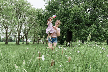 Happy mother carrying daughter with pinwheel toy while walking on grass at park - ASGF00697