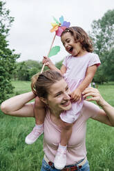 Laughing daughter holding pinwheel while sitting on mother's shoulder at park - ASGF00702