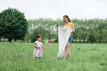Tochter mit Windrädchen in der Nähe der Mutter, die eine Decke im Park hält - ASGF00722