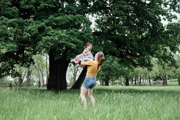 Mutter hebt ihre Tochter hoch, während sie im Park auf dem Rasen steht - ASGF00733