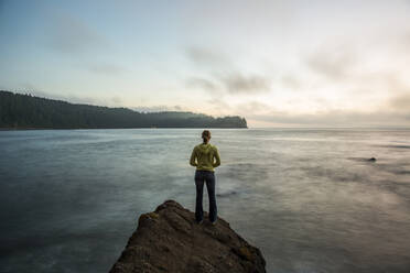 A young adult stands on the edge of a rock at sunset overlooking the Strait of Juan de Fuca, Washington. - CAVF94391