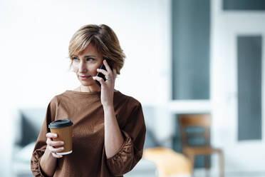 Geschäftsfrau mit Kaffeetasse, die im Büro mit einem Mobiltelefon spricht - JOSEF04934