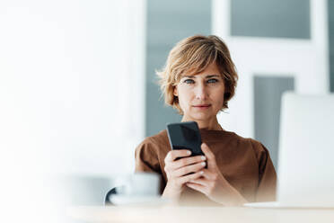 Businesswoman with mobile phone and laptop in office - JOSEF04949