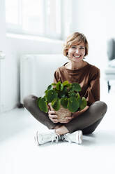 Smiling businesswoman holding potted plant while sitting cross-legged on floor - JOSEF04959