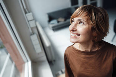 Geschäftsfrau lächelt, während sie am Fenster im Büro nach oben schaut - JOSEF04965