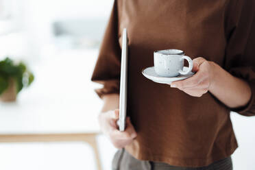 Businesswoman holding coffee cup and laptop while standing in office - JOSEF04969