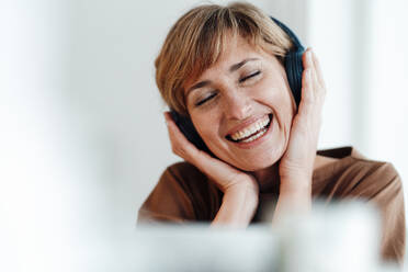 Mature businesswoman smiling while listening music through headphones in office - JOSEF04980