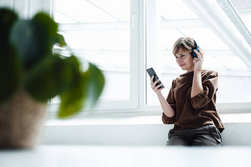 Geschäftsfrau mit Kopfhörern und Smartphone am Fenster sitzend - JOSEF04982