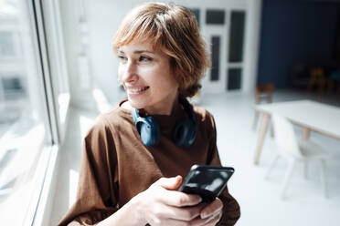 Lächelnde Geschäftsfrau mit Kopfhörern und Smartphone schaut durch ein Fenster im Büro - JOSEF04983