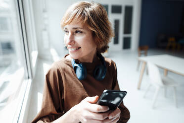 Smiling businesswoman with headphones and smart phone looking through window in office - JOSEF04983