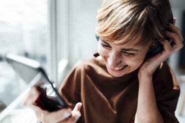 Cheerful businesswoman with mobile phone listening music through headphones in office - JOSEF04984