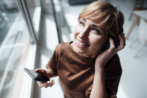 Geschäftsfrau mit Kopfhörern und Mobiltelefon am Bürofenster stehend - JOSEF04986