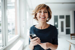 Cheerful businesswoman holding mobile phone while standing in office - JOSEF04994
