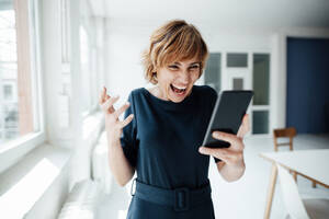 Excited businesswoman using mobile phone while standing in office - JOSEF04996