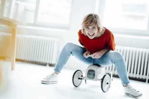 Cheerful businesswoman sitting on toy car in office - JOSEF05012