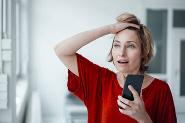 Businesswoman with mouth open and hand in hair holding smart phone in office - JOSEF05018