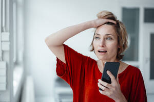Geschäftsfrau mit offenem Mund und Hand in den Haaren hält Smartphone im Büro - JOSEF05018