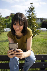 Smiling woman using smart phone while sitting on bench - GIOF13021
