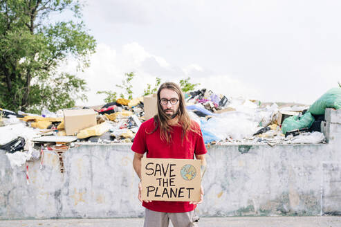 Hippie man holding cardboard with save the planet in front of near garbage dump - JCMF02008