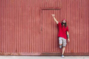 Lächelnder Hipster-Mann macht Shaka-Zeichen, während er sich an eine braune Wand lehnt - JCMF02019