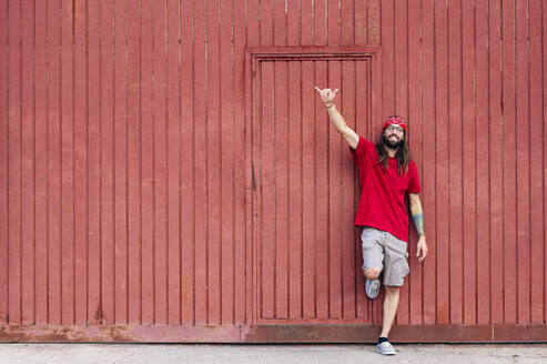 Smiling hipster man making shaka sign while leaning on brown wall - JCMF02019